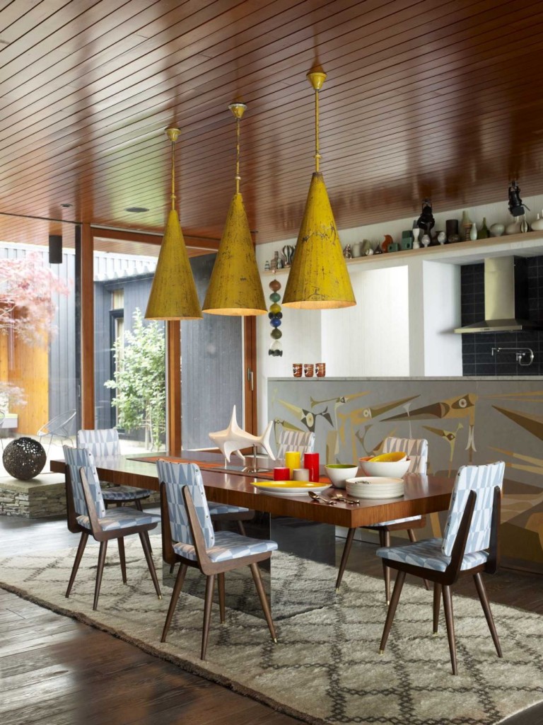 Three vintage pendant lamps from a Paris flea market hang over the dining table and echo the strongly conical shape of the fire pit’s chimney in this Shelter Island home. The dining chairs are designed by Jonathan Adler, as is the rug. Artist John Paul designed the mural depicting stylized birds that divides the kitchen from the dining area. (Credit: Courtesy of The Monacelli Press / Joshua McHugh)